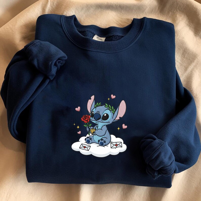 Puede incluir: Sudadera azul marino con un personaje de dibujos animados bordado sosteniendo una rosa roja. El personaje azul est&aacute; sentado en una nube con corazones y sobres. Cuello redondo y mangas largas.