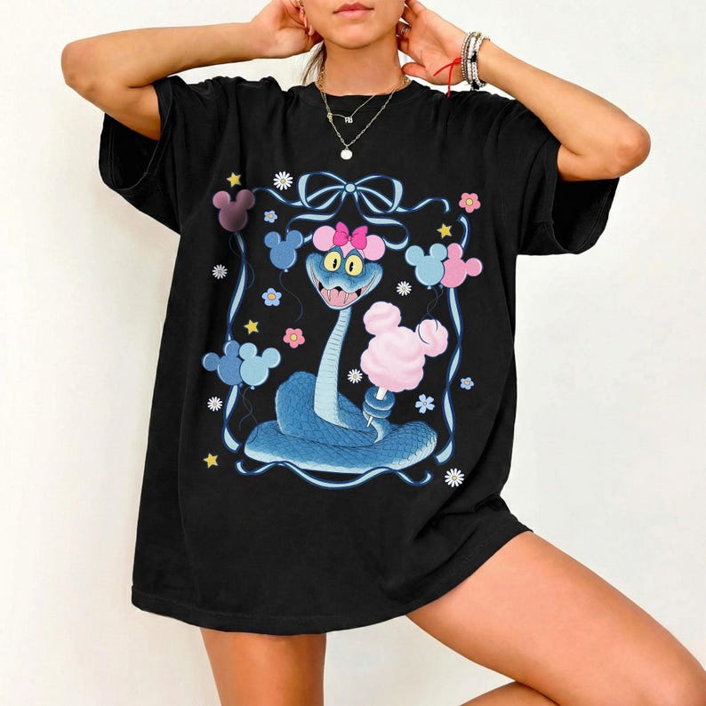 May include: Black t-shirt featuring a cartoon snake with a pink bow, holding cotton candy. The snake is blue with yellow eyes, surrounded by Mickey Mouse-shaped balloons and flowers. A whimsical design.