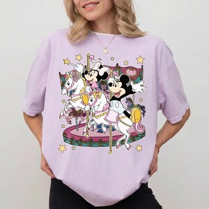 Puede incluir: Camiseta de color lila claro con un dise&ntilde;o de carrusel con los personajes de Mickey y Minnie Mouse. El carrusel tiene caballos blancos con detalles rosas y dorados, y el dise&ntilde;o incluye estrellas amarillas. La camiseta es de corte holgado.