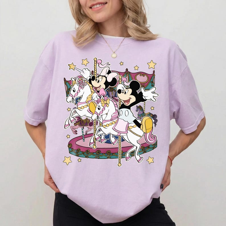 Puede incluir: Camiseta de color lila claro con un dise&ntilde;o de carrusel con los personajes de Mickey y Minnie Mouse. El carrusel tiene caballos blancos con detalles rosas y dorados, y el dise&ntilde;o incluye estrellas amarillas. La camiseta es de corte holgado.