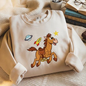 Pu&ograve; includere: Felpa beige con un cavallo dei cartoni animati ricamato, un razzo, un pianeta e una stella. Il cavallo marrone ha un naso rosa e una sella. Il razzo &egrave; rosso e giallo, e il pianeta &egrave; blu e verde.