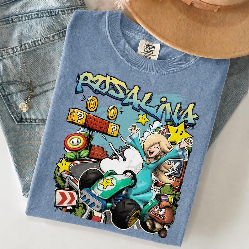 May include: A light blue t-shirt featuring a colorful graphic of Rosalina from the Mario Kart series. The design includes the word "Rosalina" in graffiti style, along with stars, coins, and a kart. The shirt is folded on top of a pair of blue jeans.