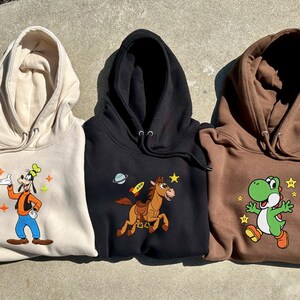 Puede incluir: Tres sudaderas con capucha en beige, negro y marr&oacute;n. Cada sudadera presenta un personaje de dibujos animados colorido: Goofy, un caballo con un cohete y Yoshi. Las sudaderas est&aacute;n hechas de un material suave.