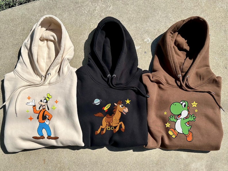 Puede incluir: Tres sudaderas con capucha en beige, negro y marr&oacute;n. Cada sudadera presenta un personaje de dibujos animados colorido: Goofy, un caballo con un cohete y Yoshi. Las sudaderas est&aacute;n hechas de un material suave.