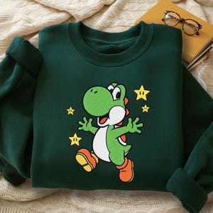 Könnte beinhalten: Dunkelgrünes Sweatshirt mit einer Cartoon-Figur von Yoshi mit orangefarbenen Stiefeln und einem weißen Bauch, umgeben von gelben Sternen. Das Sweatshirt hat einen Rundhalsausschnitt und lange Ärmel.