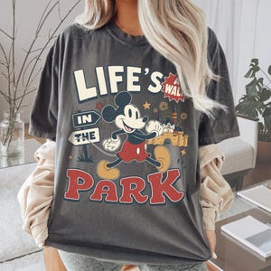 Puede incluir: Una camiseta gris carb&oacute;n de manga corta con un gr&aacute;fico de Mickey Mouse caminando. El texto dice "LIFE'S A WALK IN THE PARK" en letras negritas. El dise&ntilde;o incluye un castillo, estrellas y fuegos artificiales en colores rojo, amarillo y blanco.