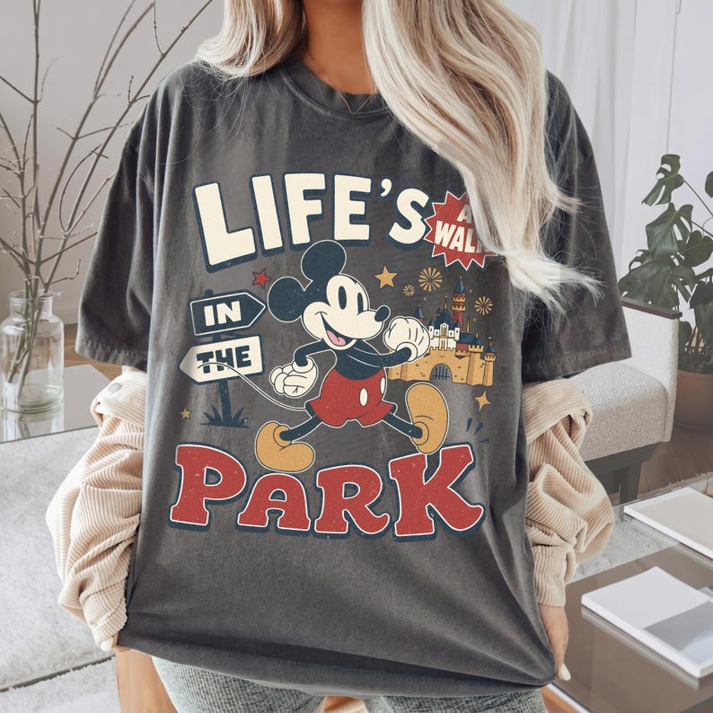 Puede incluir: Una camiseta gris carb&oacute;n de manga corta con un gr&aacute;fico de Mickey Mouse caminando. El texto dice "LIFE'S A WALK IN THE PARK" en letras negritas. El dise&ntilde;o incluye un castillo, estrellas y fuegos artificiales en colores rojo, amarillo y blanco.
