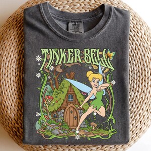 May include: Dark gray t-shirt featuring a graphic of Tinker Bell in a green dress with blue wings, in front of a fairy house. The text "TINKER BELL" is in green, with a butterfly and flowers. The shirt is on a woven surface.