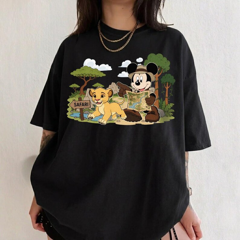 Op de afbeelding: Zwart t-shirt met een kleurrijke afbeelding van Mickey Mouse met een safarihoed, een kaart vasthoudend, en Simba het leeuwenjong. De sc&egrave;ne speelt zich af in een jungle met een "Safari"-bord.