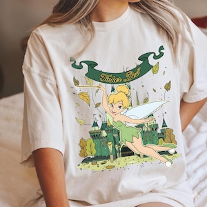 May include: Cream-colored t-shirt featuring a graphic of Tinker Bell from Peter Pan. The design includes Tinker Bell with a wand, a green castle, and the text "Tinker Bell" on a green banner. Autumn leaves are scattered around the fairy.