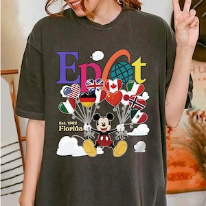 K&ouml;nnte beinhalten: Dunkelgraues T-Shirt mit Mickey Mouse, der herzf&ouml;rmige Ballons mit Flaggen h&auml;lt. Das Wort "Epcot" ist in bunten Buchstaben geschrieben, darunter steht "Florida Est. 1982". Die Ballons zeigen Flaggen verschiedener L&auml;nder.