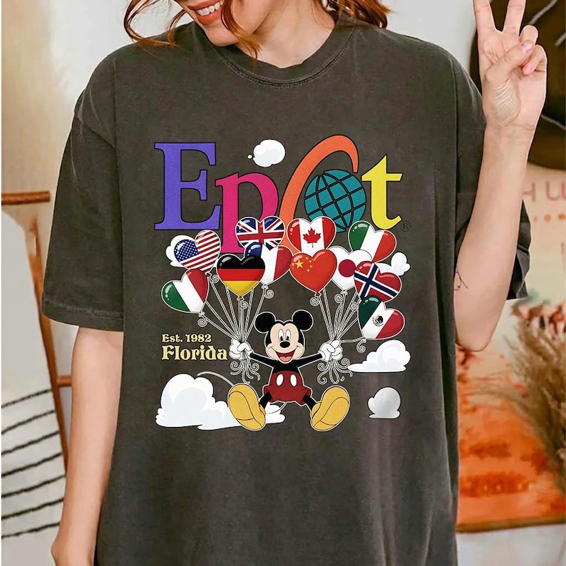 K&ouml;nnte beinhalten: Dunkelgraues T-Shirt mit Mickey Mouse, der herzf&ouml;rmige Ballons mit Flaggen h&auml;lt. Das Wort "Epcot" ist in bunten Buchstaben geschrieben, darunter steht "Florida Est. 1982". Die Ballons zeigen Flaggen verschiedener L&auml;nder.