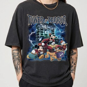 Può includere: Una t-shirt a maniche corte grigio antracite con una stampa grafica di Topolino, Pippo, Pluto e Paperino in uniforme da fattorino. Il testo Tower of Terror e Hollywood Tower è visualizzato sopra i personaggi con uno sfondo di fulmini.