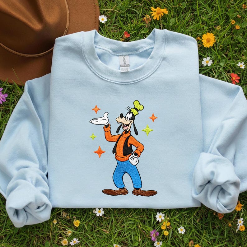 Puede incluir: Sudadera azul claro con una imagen bordada de Goofy, un personaje de dibujos animados de Disney. Goofy lleva un chaleco naranja, pantalones azules y zapatos marrones. Acentos de estrellas de colores rodean al personaje. La sudadera tiene mangas largas.