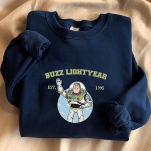 Puede incluir: Sudadera azul marino con un dise&ntilde;o de Buzz Lightyear. El dise&ntilde;o incluye el texto "BUZZ LIGHTYEAR" encima de una ilustraci&oacute;n del personaje, con "EST. 1995" debajo. Tiene cuello redondo y pu&ntilde;os acanalados.