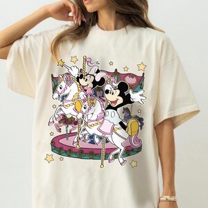 Puede incluir: Camiseta color crema con un dise&ntilde;o de carrusel colorido con Mickey Mouse, Minnie Mouse y otros personajes. El carrusel tiene caballos blancos, detalles en rosa y morado y estrellas doradas. El dise&ntilde;o es caprichoso y juguet&oacute;n.