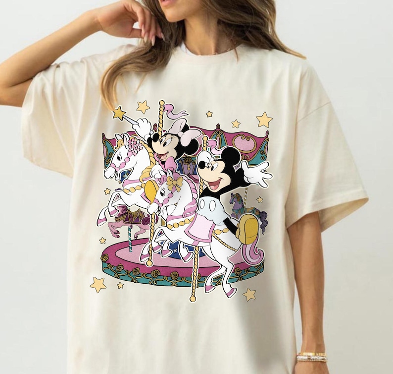 Puede incluir: Camiseta color crema con un dise&ntilde;o de carrusel colorido con Mickey Mouse, Minnie Mouse y otros personajes. El carrusel tiene caballos blancos, detalles en rosa y morado y estrellas doradas. El dise&ntilde;o es caprichoso y juguet&oacute;n.