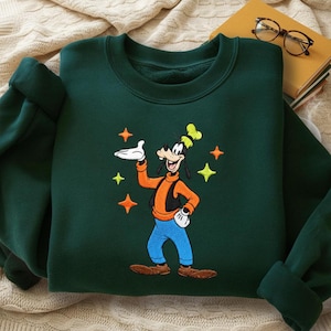 Puede incluir: Sudadera verde oscuro con una imagen bordada de Goofy, un personaje de dibujos animados de Disney. Goofy lleva una camisa naranja, pantalones azules y un sombrero amarillo. El dise&ntilde;o incluye estrellas amarillas y naranjas. La sudadera tiene cuello redondo y mangas largas.