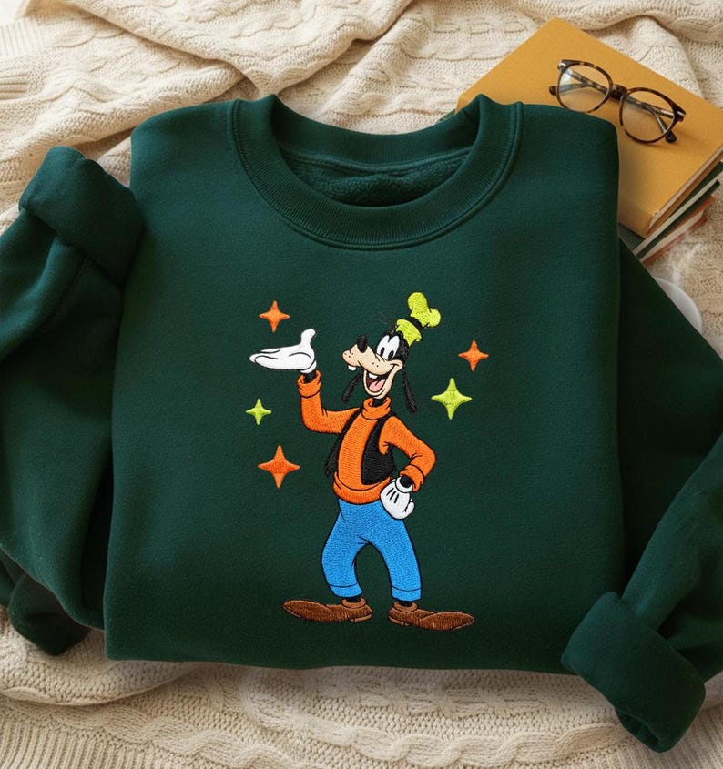 Puede incluir: Sudadera verde oscuro con una imagen bordada de Goofy, un personaje de dibujos animados de Disney. Goofy lleva una camisa naranja, pantalones azules y un sombrero amarillo. El dise&ntilde;o incluye estrellas amarillas y naranjas. La sudadera tiene cuello redondo y mangas largas.