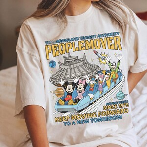 May include: Cream-colored t-shirt featuring a colorful graphic of the PeopleMover ride with Mickey Mouse, Minnie Mouse, Donald Duck, Daisy Duck, and Goofy. The shirt includes the text "TOMORROWLAND TRANSIT AUTHORITY PEOPLEMOVER" and "KEEP MOVING FORWARD TO A NEW TOMORROW."