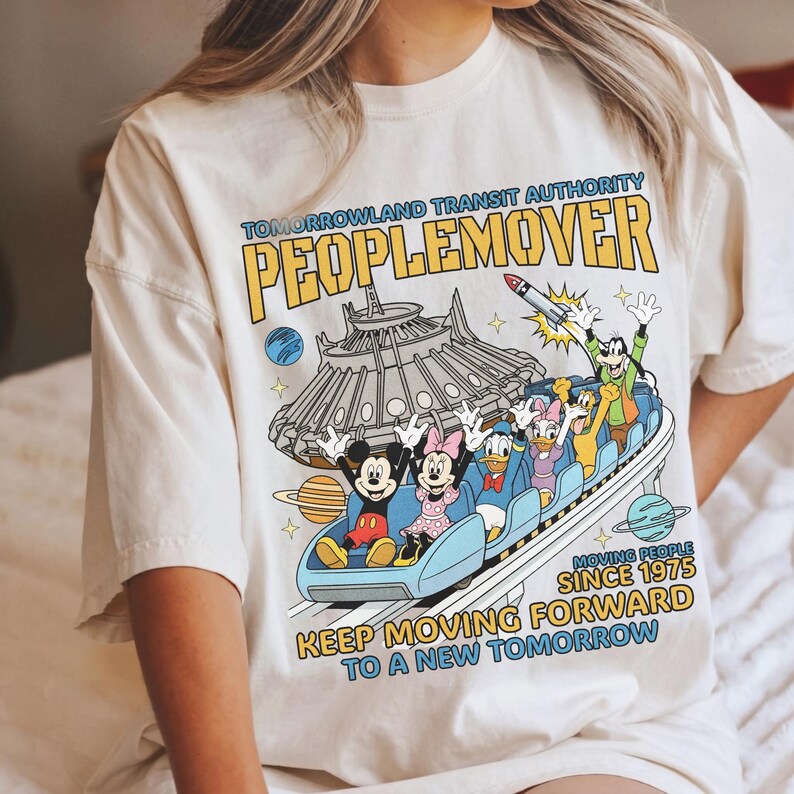 May include: Cream-colored t-shirt featuring a colorful graphic of the PeopleMover ride with Mickey Mouse, Minnie Mouse, Donald Duck, Daisy Duck, and Goofy. The shirt includes the text "TOMORROWLAND TRANSIT AUTHORITY PEOPLEMOVER" and "KEEP MOVING FORWARD TO A NEW TOMORROW."