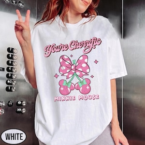 Pode incluir: Camiseta branca com um la&ccedil;o rosa com bolinhas brancas e dois pares de cerejas rosas com talos verdes. O texto "You're Cherriffic" e "MINNIE MOUSE" &eacute; impresso acima e abaixo do gr&aacute;fico em rosa.