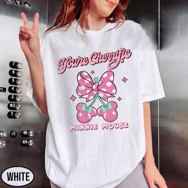 Pode incluir: Camiseta branca com um la&ccedil;o rosa com bolinhas brancas e dois pares de cerejas rosas com talos verdes. O texto "You're Cherriffic" e "MINNIE MOUSE" &eacute; impresso acima e abaixo do gr&aacute;fico em rosa.
