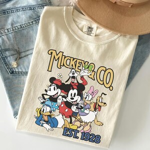 May include: Cream-colored t-shirt featuring a graphic of Mickey Mouse and friends, with the text "Mickey & Co." and "EST. 1928" in a vintage style. The shirt is folded on top of blue jeans.