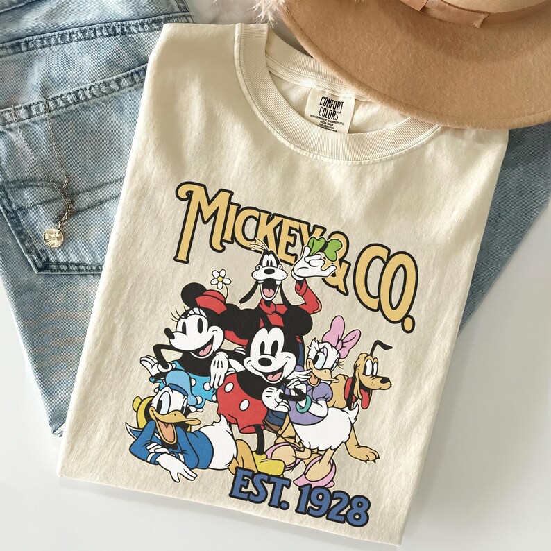 May include: Cream-colored t-shirt featuring a graphic of Mickey Mouse and friends, with the text "Mickey & Co." and "EST. 1928" in a vintage style. The shirt is folded on top of blue jeans.