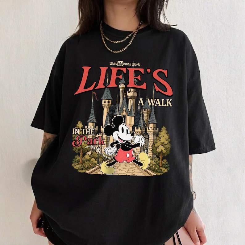 Puede incluir: Camiseta negra con un gr&aacute;fico de Mickey Mouse caminando frente a un castillo, con el texto "LIFE'S A WALK IN THE PARK" y "Walt Disney World" arriba. El dise&ntilde;o es en rojo, amarillo y beige.