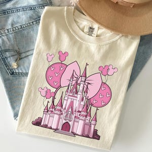 May include: Cream-colored t-shirt featuring a pink castle graphic with a large bow and Mickey Mouse balloon accents. The shirt is folded on top of blue jeans and a tan hat.