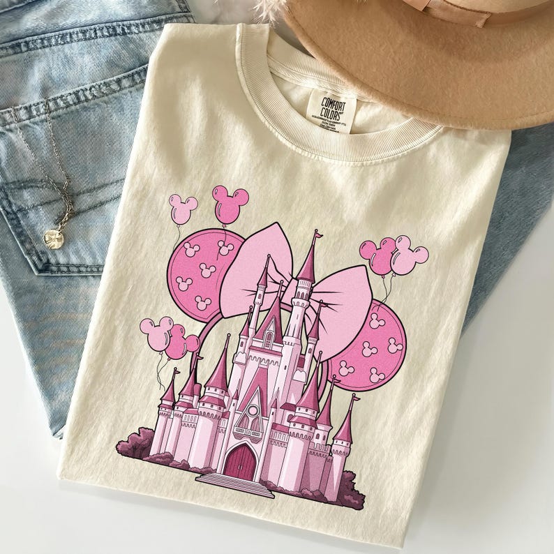 May include: Cream-colored t-shirt featuring a pink castle graphic with a large bow and Mickey Mouse balloon accents. The shirt is folded on top of blue jeans and a tan hat.