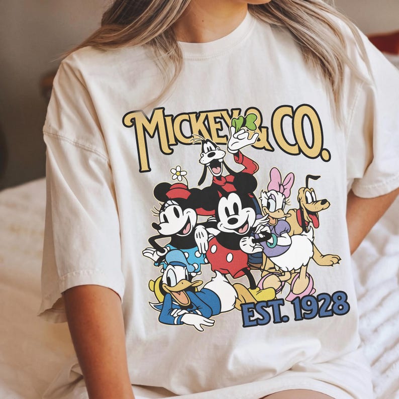 May include: Cream-colored t-shirt featuring a graphic of Mickey Mouse and friends, including Minnie Mouse, Donald Duck, and Goofy. The design includes the text "Mickey & Co." and "EST. 1928" in a vintage style.