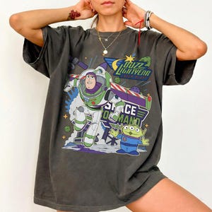 Op de afbeelding: Donkergrijs T-shirt met een afbeelding van Buzz Lightyear en een alien uit de film Toy Story. De afbeelding bevat de tekst "Buzz Lightyear Laser Blast" en "Space Command". Het ontwerp is in groen, paars, blauw en wit.