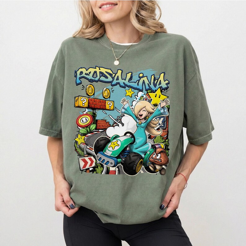 May include: Sage green t-shirt featuring a colorful graphic of Rosalina from Mario Kart. The design includes the word "Rosalina" in graffiti style, along with stars, coins, and a kart. The shirt is a comfortable, oversized fit.