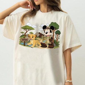 Op de afbeelding: Cr&egrave;mekleurig T-shirt met een cartoon safarisc&egrave;ne. Mickey Mouse, met een safarihoed, wijst naar een kaart naast een leeuwenwelp. De sc&egrave;ne bevat bomen, wolken en een "Safari"-bord.