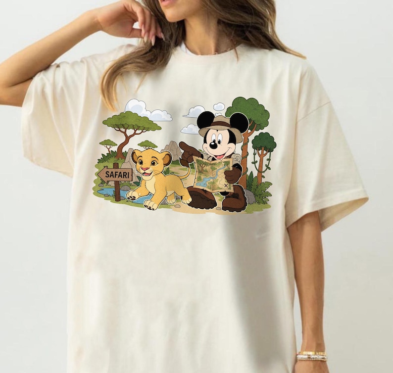 Op de afbeelding: Cr&egrave;mekleurig T-shirt met een cartoon safarisc&egrave;ne. Mickey Mouse, met een safarihoed, wijst naar een kaart naast een leeuwenwelp. De sc&egrave;ne bevat bomen, wolken en een "Safari"-bord.