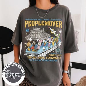 May include: Dark gray t-shirt featuring a graphic of the PeopleMover ride with Mickey, Minnie, Donald, and other Disney characters. The shirt has the text "Tomorrowland Transit Authority PeopleMover" and "Moving People Since 1975".