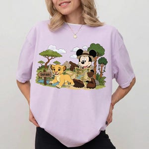 Op de afbeelding: Lichtpaars T-shirt met een cartoon illustratie van Mickey Mouse in safari-outfit, wijzend naar een kaart, en Simba uit The Lion King. Een bord leest "Safari".