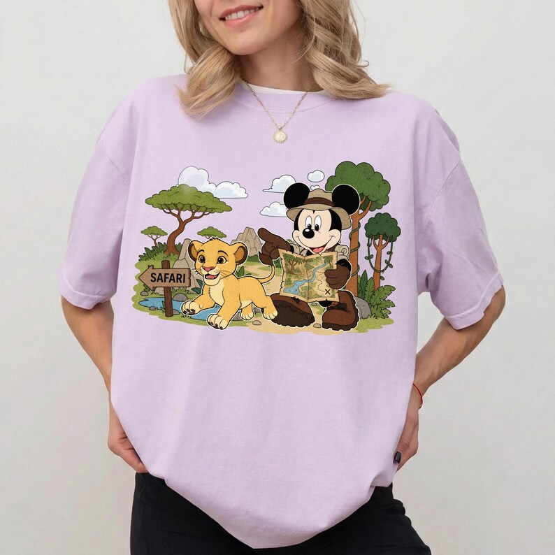 Op de afbeelding: Lichtpaars T-shirt met een cartoon illustratie van Mickey Mouse in safari-outfit, wijzend naar een kaart, en Simba uit The Lion King. Een bord leest "Safari".