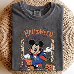 May include: A charcoal gray crew neck t-shirt featuring a graphic of Mickey Mouse dressed as a vampire. The design includes the text HALLOWEEN PARTY, pumpkins, a purple witch hat, a skull, a cauldron, and bare tree branches in a frame.