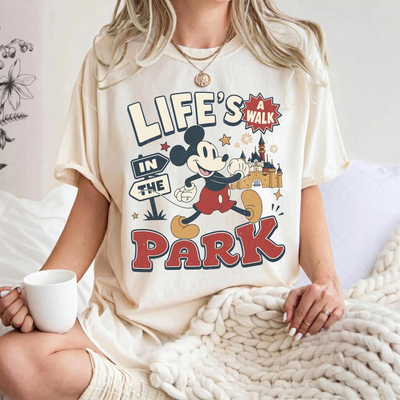 Puede incluir: Una camiseta de color crema con un gr&aacute;fico de Mickey Mouse caminando. El texto dice "LIFE'S A WALK IN THE PARK" junto a una ilustraci&oacute;n de un castillo, estrellas y fuegos artificiales. El dise&ntilde;o utiliza colores rojo, amarillo y azul.