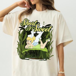 May include: Cream-colored t-shirt featuring a cartoon illustration of a fairy lying on a leaf, surrounded by green foliage. The text "Short N Sweet" is above the fairy. The fairy has blonde hair, green clothing, and blue wings.