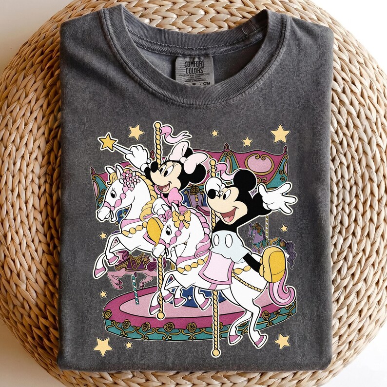 Puede incluir: Camiseta gris oscuro con un dise&ntilde;o de carrusel con Mickey y Minnie Mouse. La imagen incluye caballos blancos, detalles rosas y amarillos, y estrellas doradas. La camiseta tiene cuello redondo.