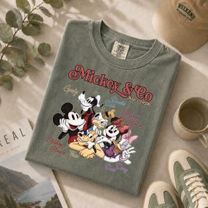 May include: A sage green t-shirt featuring a graphic of Mickey Mouse and friends, with the text "Mickey & Co Since 1928". The shirt is surrounded by a beige cap, a mug, and sneakers.