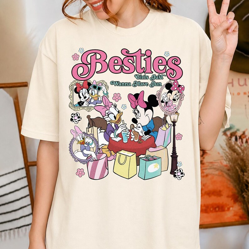 May include: Cream-colored t-shirt featuring a graphic of Minnie Mouse, Daisy Duck, and other Disney characters. The design includes the text "Besties Girls Just Wanna Have Fun" in pink lettering, along with floral accents and shopping bags.