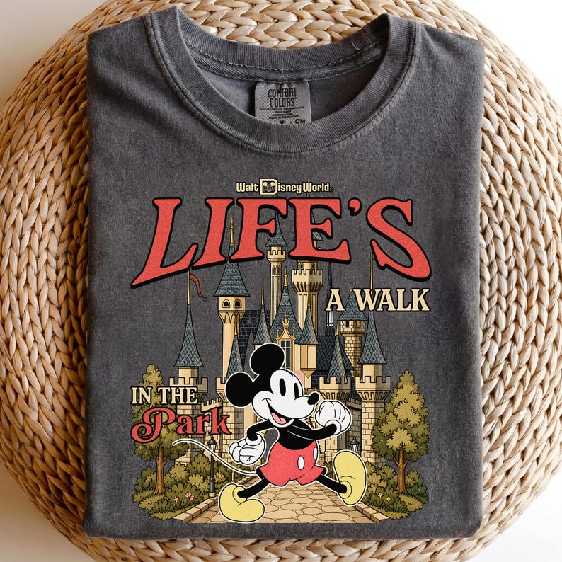 Puede incluir: Camiseta gris oscuro con un gr&aacute;fico de Mickey Mouse caminando frente a un castillo y el texto "Life's a Walk in the Park" en rojo y dorado. La camiseta es de la marca Comfort Colors.