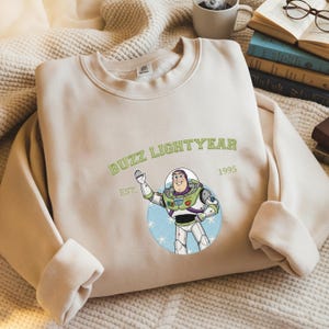 Puede incluir: Sudadera beige con un dise&ntilde;o de Buzz Lightyear. El dise&ntilde;o incluye el texto "BUZZ LIGHTYEAR" sobre una ilustraci&oacute;n del personaje, con "EST. 1995" debajo. El dise&ntilde;o es en tonos verdes, blancos y azules.