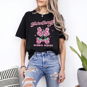 Pode incluir: Camiseta preta com la&ccedil;o rosa de bolinhas brancas e design de cereja da Minnie Mouse. O texto "You're Cherrifi" e "MINNIE MOUSE" est&atilde;o impressos na camiseta. A modelo est&aacute; usando jeans azuis.