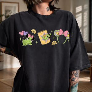 Peut inclure: T-shirt noir avec des graphiques de dessins animés représentant un caméléon vert avec un nœud rose, un livre avec un caméléon et des oreilles de souris avec un nœud rose. Les graphiques sont verts, roses, jaunes et violets.